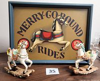 Signed Merry-Go-Round Rides wood picture with a blue frame and two Victorian style rocking horse figurines in pastel colors on either side.