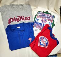 Four folded Phillies shirts displayed: grey Phillies shirt, white 1993 National League East Champions shirt, blue sleeveless zip shirt with 'P', red raglan sleeve shirt with Phillies applique.
