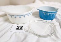 Two vintage Pyrex casserole dishes, one white with blue garland pattern and one blue with white snowflake garland pattern, along with a clear glass lid.