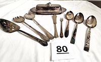 Photo showing nine silver-plated utensils including various spoons, fork, spreader, and butter dish with floral handle