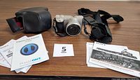 Full lot overview showing camera with strap attached, leather camera case, and several informational pamphlets spread on a wooden table surface.