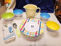 Eight pastel plaid melamine dinner plates stacked with four colorful melamine bowls (yellow, green, two blue), light yellow Oneida Petals pitcher, seashore-themed kitchen towel, and ice cream scoop with pale green handle arranged on white surface