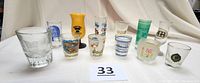 Full view of all twelve assorted beach and travel theme shot glasses on white table, showing variety of colors, logos and sizes