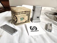 Three silver tone lighters and ceramic coin bank resembling $10 bill stack on white background with lot number card 69