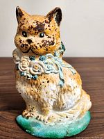 Front view showing painted tabby cat with blue ribbon and white flowers around the neck, paint loss and spots of rust visible.