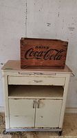 Wooden Coca Cola crate resting on the top of the metal utility table, showing the Coca Cola branding on the crate's side and the drawer and cabinet of the table.
