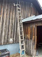 Aluminum extension ladder leaning against an exterior wooden wall near a shed roof. The ladder shows dirt and weathering, indicating outdoor use.