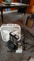White box fan, two small portable heaters in front of wooden furniture.