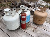 Four propane or gas cans and one red fire extinguisher on an outdoor wooden surface, with visible signs of wear and rust on the tanks.