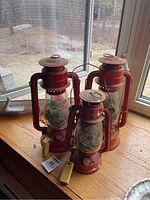 Three red metal railway lanterns with glass chimneys on a windowsill, containing artificial greenery, showing overall condition and tags.