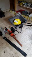 Wide view of electric trimmer, yellow hard hat, and leaf blower on floor indoors.