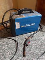 Blue Mastercraft welding machine with power cord and welding gun visible on carpet.