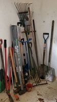 Photo showing multiple garden tools including shovels, rakes, and hand tools bundled together and leaning against a wall.