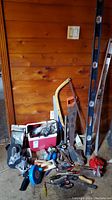 Wide shot showing assorted hand tools including several levels, bow saw, red cooler, axes, wrenches, and red clamps against a wooden wall.