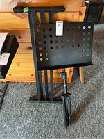 Black metal music stand in folded position leaning against wooden furniture on carpeted floor.
