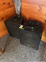 Two Yorkville Performance Series speakers side by side on carpeted floor against wooden wall, cables coiled on top