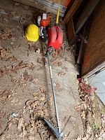 Full view of the gas-powered weedeater alongside the yellow hard hat, protective goggles, and red gas can on a floor with dried leaves.