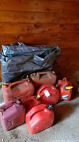 Various red and dark red gasoline jerry cans arranged on wooden floor with a folded green tarp behind them. Some have yellow spouts attached. Show signs of dirt and wear.