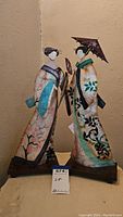 Full view of two Geisha figures on wooden bases, dressed in traditional attire with floral decorations and holding fan and parasol.