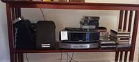 Wide view of shelf showing Bose unit, speakers, remote, CD cartridges, storage case and CDs.