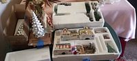 Wide shot of Christmas village figurines and pieces packed in foam trays on a table, including an angel figurine in background and a silver metallic tree.