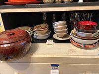 Photo showing the entire dishware set with a prominent large brown covered serving dish, several stacks of various sizes of ceramic bowls, saucers, and multiple clear glasses arranged on shelves.
