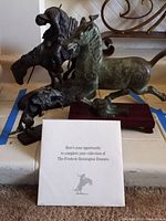Two bronze sculptures with a white paper showing collection opportunity placed in front. Horse sculpture on wooden base and cowboy on horseback figure visible.