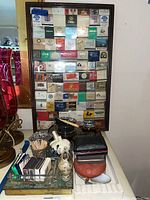 Framed collection of matchbooks displayed on a wall, with a table underneath holding various smoking-related items including ashtrays and novelty pieces.