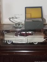 Side view of beige 1932 Cadillac model car inside clear display case with hood ornaments and box in background