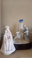 Two porcelain figure music boxes, one Madonna and Child in white finish and one Asian lady with attendants in pastel colors on wooden base.