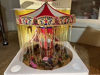 Close-up of the carousel showing red and white canopy, gold detailed center pillar, and hanging swing seats with miniature figures seated.