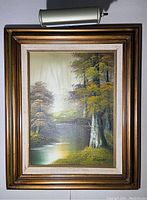 Framed signed oil painting with lamp above it, showing full artwork and frame. Painting depicts a landscape with trees and a water body.