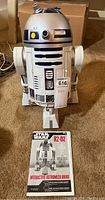 Front view of R2-D2 droid standing next to original box and manual on carpet.