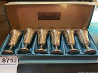Six sterling silver cordial cups arranged in a lined box with 'Sterling Silver' plaque on inside lid. The box interior is beige with blue satin fabric lining on bottom.