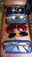 Five pairs of glasses and sunglasses arranged vertically on a wooden surface with some cases visible, showing different frame styles and lens colors.