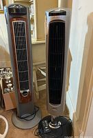 Two tower fans, one Lasko with wood grain accents and one DeLonghi, showing their front and side views.