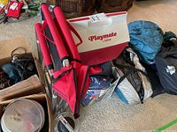 Camping gear including red folding chair, Igloo Playmate cooler, tents in bags on carpet floor