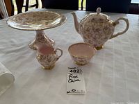 Full set displayed on a white tablecloth showing a pedestal cake stand, teapot with lid, creamer, and sugar bowl with a floral and gold pattern.