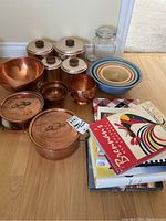 Copperware pots, bowls, canisters, glass jar, Pyrex mixing bowls, and cookbooks arranged on floor.