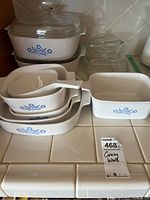 Photo showing multiple CorningWare dishes stacked including oval, rectangular, and bowl shaped dishes with blue cornflower design and a glass lid in background