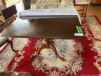 Full view of dining table on floral carpet, wooden rectangular top with turned pedestal bases and three curved feet.