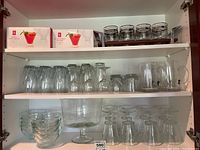 Shelf with various styles of clear glasses, mugs, a large pedestal bowl, and two boxed sets of PC glass candle holders on the top shelf.