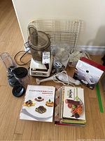 Overview of all kitchen appliances and cookbooks in the lot, showing food processors, hand mixer, immersion blender, can opener box, and cookbooks on wood floor.