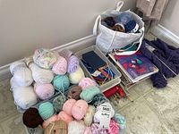 Overall collection of yarn skeins, knitting needles, and patterns arranged on floor with a white bag containing more yarn.