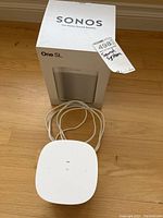 Photo of Sonos One SL product box with white wireless controller device and attached power cable placed in front on a hardwood floor.