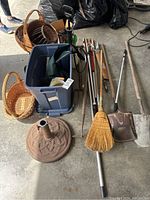 Photo showing garden tools such as shovels, broom, rakes, a large blue plastic bin and a terracotta umbrella stand base on concrete floor.