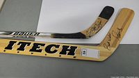Two autographed hockey sticks lying side by side; one is a Bauer Supreme goalie stick with an autograph on the blade, the other is a beige Sher-Wood ITECH Profile stick with an autograph on the shaft.