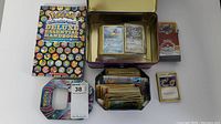 Photo showing the Pokemon Deluxe Essential Handbook book, open tin lunchbox with Pokemon cards inside, and a box of additional cards.