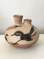 Front angle view showing the pottery vase with two necks, painted bird and feather design in black and brown on natural tan body, terracotta color rims and base band.