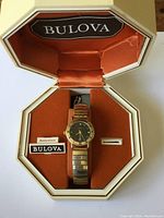 Vintage Bulova wristwatch inside original box with orange lining, showing watch face and metal expandable bracelet.